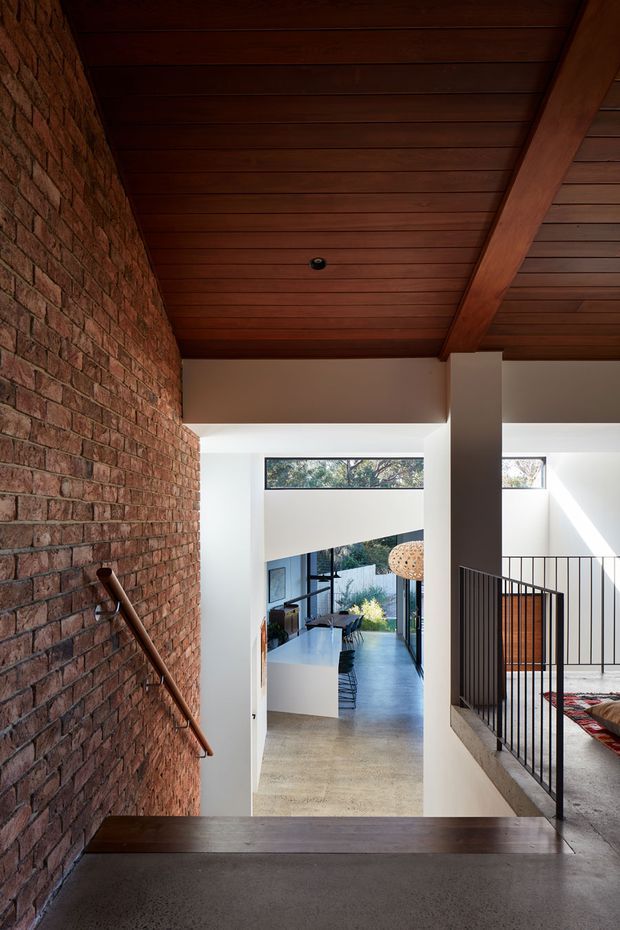 Interior view of a contemporary multi-level home hallway and stair landing. A raw exposed brick wall with timber handrail runs along the left, while a richly stained timber-planked ceiling with substantial exposed beam spans overhead. White plaster walls, a polished concrete floor, black metal balustrade, and a stair opening frame a bright kitchen and living area beyond. The space has strong natural light and a warm, material-focused architectural character.