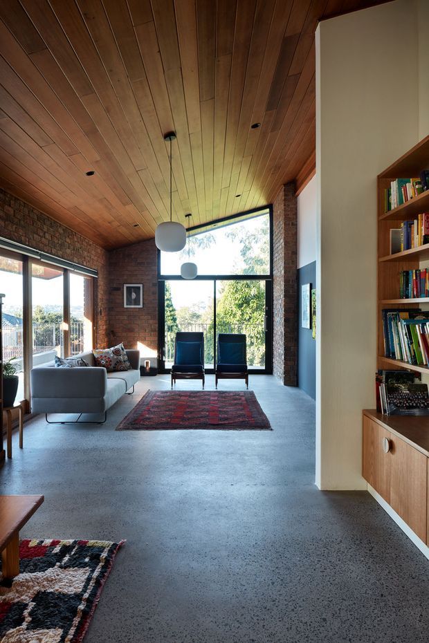 A warm, open-plan living room with exposed red brick walls, a rich timber-lined ceiling, polished grey concrete flooring, and large black-framed windows and glazed doors overlooking greenery. The room contains a pale sofa, dark lounge chairs, pendant lights, patterned rugs, and a built-in timber bookshelf and cabinetry along the right wall.