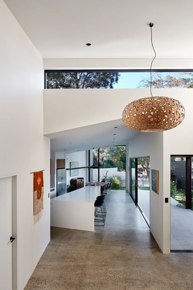 A bright, contemporary open-plan home interior viewed from an entry or hallway. The space features polished concrete flooring, white walls and ceilings, a large white kitchen island with dark bar stools, extensive black-framed glazing, and a sculptural woven pendant light. A living and dining area extends beyond the kitchen, with a garden visible through the windows and doors.