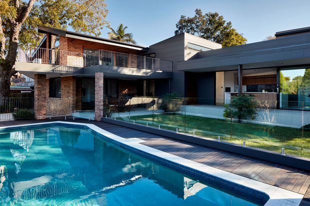 Exterior view of a contemporary multi-level residence with strong horizontal rooflines, dark grey rendered volumes, exposed red brick walls, extensive glazing and balconies. A large swimming pool with glass safety fencing and timber decking occupies the foreground, with landscaped garden areas surrounding the house.