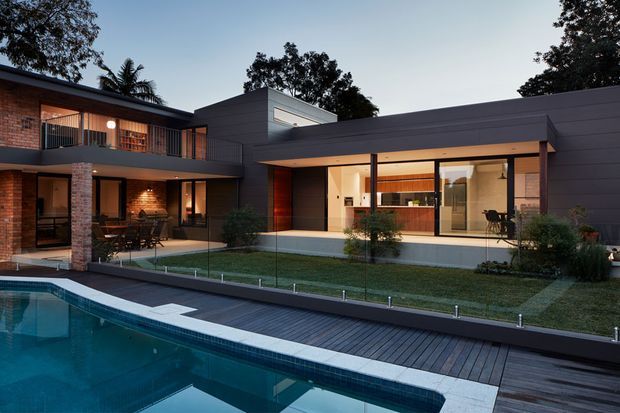 Contemporary two-storey residence arranged around a landscaped courtyard and swimming pool. The home combines dark horizontal cladding, exposed red brick, extensive glazing and warm timber details, with a covered outdoor entertaining area overlooking the pool.