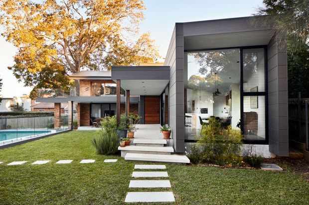 Exterior view of a contemporary multi-level residence with a flat-roofed, box-like extension featuring expansive floor-to-ceiling glazing. The home combines dark grey cladding, warm timber and exposed brick elements, with a landscaped lawn, stepping-stone path, covered entry, and swimming pool visible to the side.