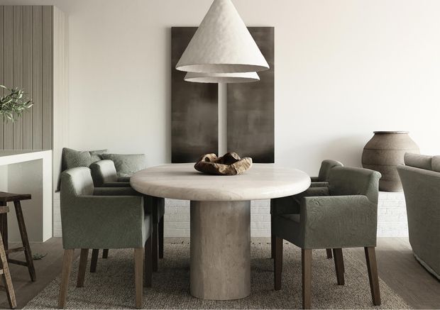 A refined dining area with a round stone-look pedestal table surrounded by upholstered dining chairs. A large sculptural conical pendant hangs centrally above the table, while a textured neutral rug, white-painted brick wall, vertical timber wall panelling, dark artwork, and a ceramic vessel create a warm, tonal interior.