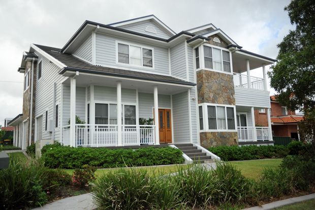 Exterior view of a large multi-storey suburban home with pale horizontal weatherboard cladding, steep gabled roof sections, covered verandas and balconies, white railings and trims, timber entry door, and a prominent natural stone facade feature. The house is set behind landscaped garden beds and a footpath.