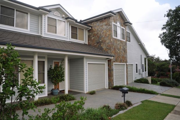 Exterior view of a multi-storey suburban residential building with grey horizontal weatherboard cladding, a prominent natural irregular-stone-clad central gable, dark shingle-style roofing, white-trimmed windows, covered entry porch, and multiple integrated garage doors. Concrete driveways, a footpath, lawn, and landscaped planting are visible in the foreground.