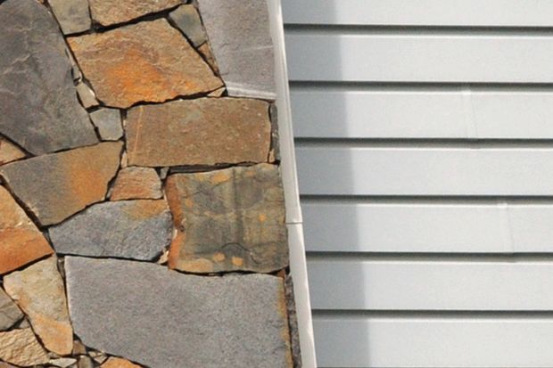 Close-up comparison of two exterior wall finishes: irregular natural stone veneer with warm earthy tones on the left and pale horizontal metal or weatherboard-style cladding on the right, separated by a narrow trim.