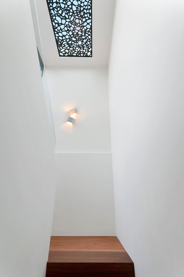 A narrow, minimalist stairway viewed looking upward, with dark timber stair treads enclosed by smooth white plaster walls. A geometric wall light illuminates the landing, and a decorative patterned skylight or ceiling panel provides a distinctive overhead feature.