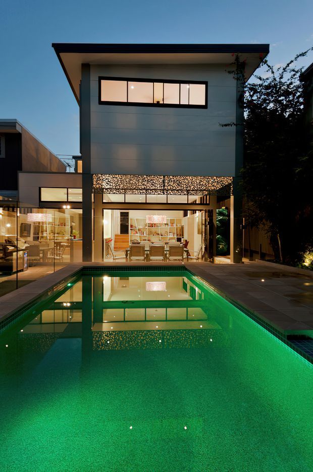 Twilight exterior view of a contemporary multi-storey residence with a flat roof, large glazed openings, and a decorative patterned screen across the upper terrace or facade. A long rectangular swimming pool dominates the foreground, reflecting the illuminated house. The open-plan living and dining area is visible through the ground-floor glazing, with tiled paving and landscaped planting around the pool.