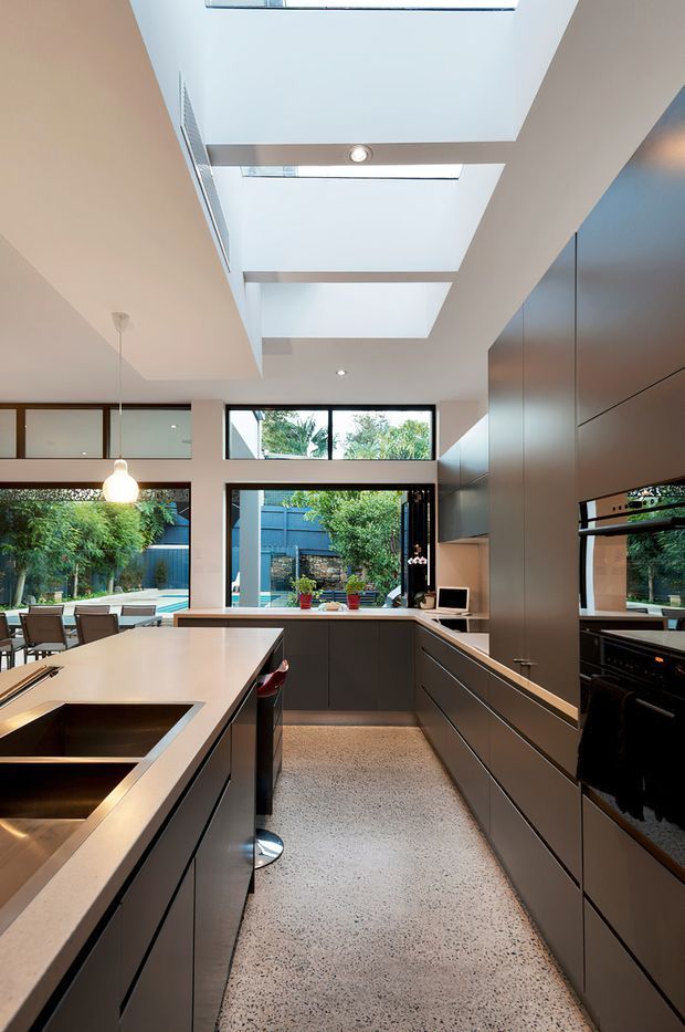 Contemporary galley-style kitchen with a large central island, integrated dark cabinetry, pale stone benchtops, black undermount sinks, and extensive glazing to a planted outdoor courtyard. Multiple skylights and a high ceiling bring abundant natural light into the space. A dining area is visible beyond the kitchen.
