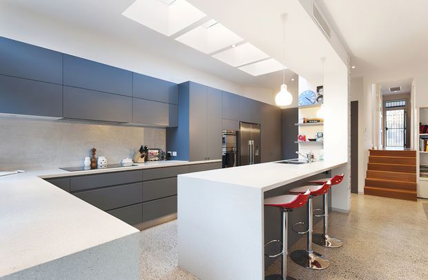 Bright contemporary open-plan kitchen with extensive matte dark grey-blue cabinetry, integrated stainless-steel appliances, pale stone benchtops, and a large white waterfall island with red bar stools. Skylights provide strong natural light, while a stairway and entry area are visible beyond the kitchen.