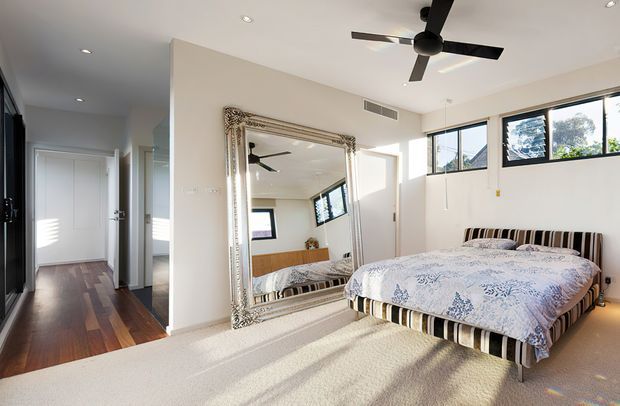 Bright contemporary bedroom with a large upholstered bed, striped bed base and patterned bedding. A substantial ornate framed mirror leans against the wall, reflecting the bed and ceiling fan. The room has pale walls, light carpet, dark timber flooring in the adjacent hallway, high horizontal windows, recessed ceiling lights and a black ceiling fan. A doorway and corridor are visible to the left.