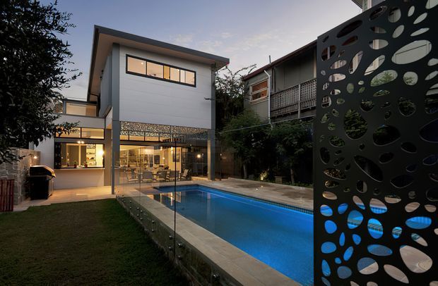 Twilight view of a contemporary multi-level home with a white rendered upper storey, extensive ground-floor glazing, and a rectangular swimming pool. A sculptural perforated dark privacy screen frames the foreground, while a timber-clad neighbouring residence and landscaped garden sit behind the pool.