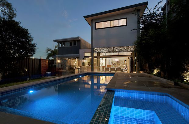 Twilight exterior view of a contemporary multi-level residence opening onto a large illuminated swimming pool and raised spa. The home has clean rectilinear forms, a projecting low-pitched roof, dark-framed glazing, a pale rendered facade, and patterned screening above the glazed ground-floor living area. Dense landscaping and boundary fencing frame the pool terrace.