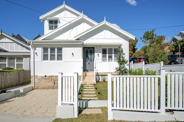 Front exterior view of a compact white multi-gabled house with weatherboard-style cladding, decorative gable detailing, louvred windows, a small covered entry porch, front lawn, paved driveway, and white picket-style fencing. The sunny suburban facade has a coastal traditional character.