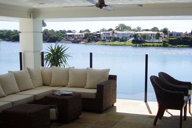A covered waterfront outdoor living area with a woven wicker sectional sofa, matching armchairs and coffee table, tiled flooring, ceiling fan, and a glass balustrade overlooking calm water and houses on the opposite shore.