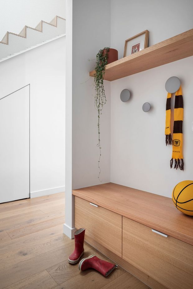 Bright contemporary entryway with pale timber flooring and built-in light-wood cabinetry. A long timber shelf displays framed art and a trailing plant, while round wall-mounted fixtures and a yellow-and-black sports scarf add visual interest. Red boots and a basketball sit near the low storage unit. The adjacent doorway and upper perforated stair guard are visible at left.