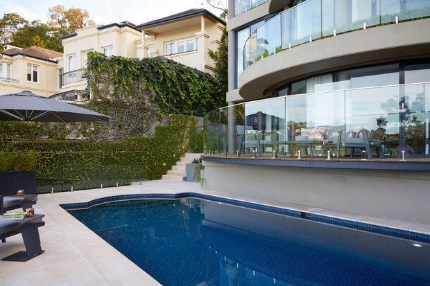 A landscaped residential pool courtyard beside a contemporary multi-level home. The deep blue tiled swimming pool is bordered by pale stone paving, outdoor lounge furniture and umbrellas, with glass balustrades, expansive glazing and curved balconies overlooking the pool. A neighbouring traditional-style residence is partially visible behind a tall planted boundary wall.