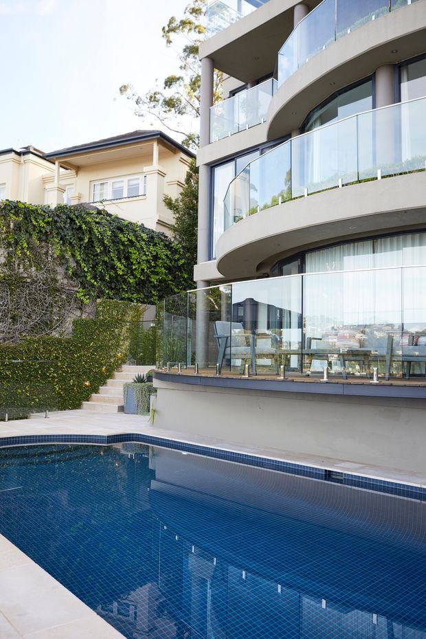 Exterior architectural photograph of a contemporary multi-storey residence beside a tiled swimming pool. The building features curved concrete balcony edges, extensive floor-to-ceiling glazing, frameless glass balustrades, and a pale rendered facade. A landscaped ivy-covered retaining wall and steps lead down to the pool, while a more traditional neighbouring house is visible behind.