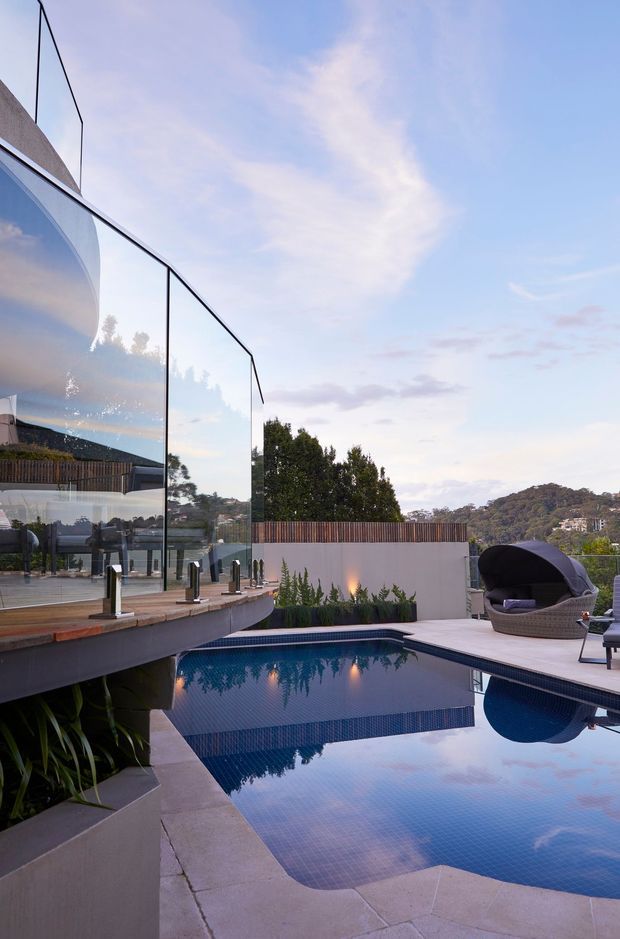 Contemporary residential pool area with a deep blue tiled swimming pool, pale stone paving, raised timber deck, frameless glass balustrades, and an outdoor lounge chair. The scene overlooks a leafy suburban landscape and is photographed at dusk, with reflections of the sky and surrounding vegetation on the water.