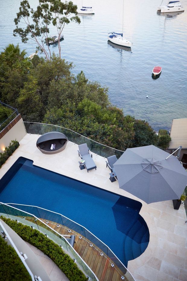 Elevated view of a landscaped waterfront terrace with a large blue swimming pool, pale stone paving, glass balustrades, lounge chairs, a fire pit, and a large grey umbrella. Boats are visible on the calm water beyond the garden.
