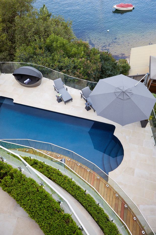 Elevated view of a landscaped outdoor pool area beside a clear waterfront, featuring a curved dark-blue swimming pool, pale stone paving, timber decking, glass balustrades, sun loungers, a large grey umbrella, and dense planted terraces. A small boat is visible on the water beyond the property.