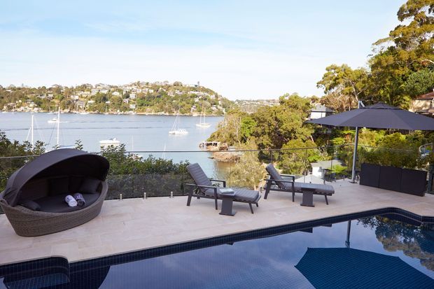 A landscaped waterfront outdoor terrace with a swimming pool overlooking a sheltered bay. The pale stone-paved deck includes sculptural outdoor loungers, a rounded covered daybed, dark umbrellas, screening planters, and a glass balustrade, with boats and tree-covered residences visible across the water.