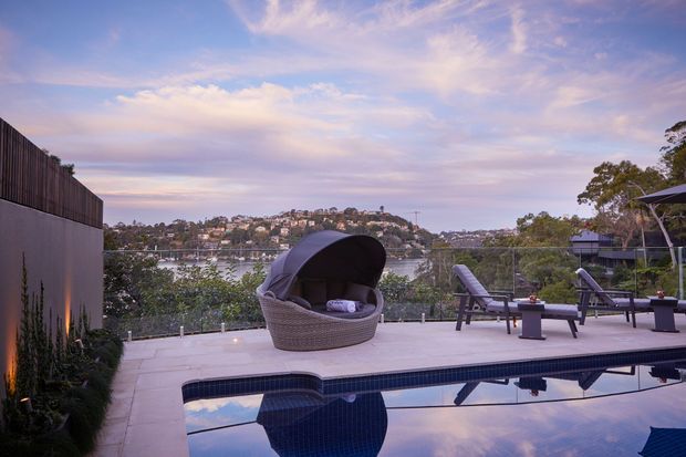 Outdoor pool terrace overlooking a calm waterfront inlet, with a large woven daybed, lounge chairs, a tiled pool, glass balustrade and landscaped screening. The scene is photographed at dusk with a soft pastel sky and residential hillside beyond.