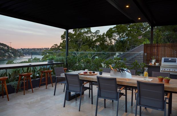 Covered outdoor entertaining terrace with a large timber dining table, bar seating, built-in outdoor kitchen and barbecue, glass balustrade, and views across a leafy waterfront suburb. The space is furnished in a contemporary style with a dark overhead roof structure and warm evening light.