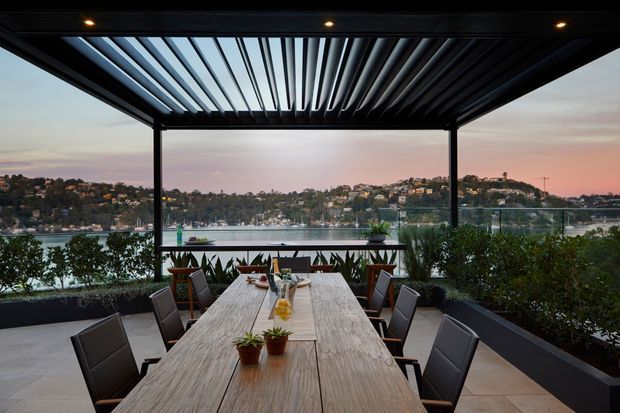 A contemporary outdoor dining terrace overlooking a calm waterfront, with a long timber dining table, dark chairs, raised planter beds, and a black steel pergola featuring adjustable louvres and integrated spotlights. The scene has an expansive harbour view at dusk.