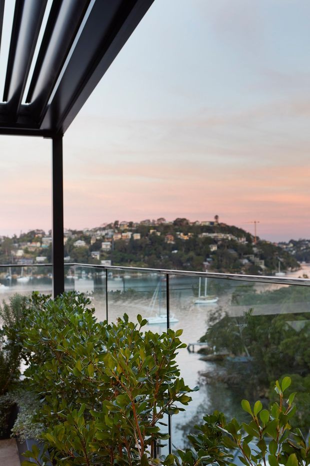 A covered balcony or terrace with dark metal framing and clear glass balustrades, overlooking a calm waterway and a densely built, tree-covered hillside at dusk. Large glossy-leaved plants occupy the foreground, while the roof structure and railing frame the scenic waterfront outlook.