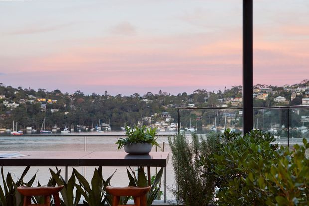 A balcony or terrace overlooking a calm waterfront inlet, photographed at dusk. The space features a dark timber or metal-framed outdoor dining table, two terracotta-toned stools, potted plants, glass balustrading, and a large dark vertical frame, with residential hills and moored boats across the water.