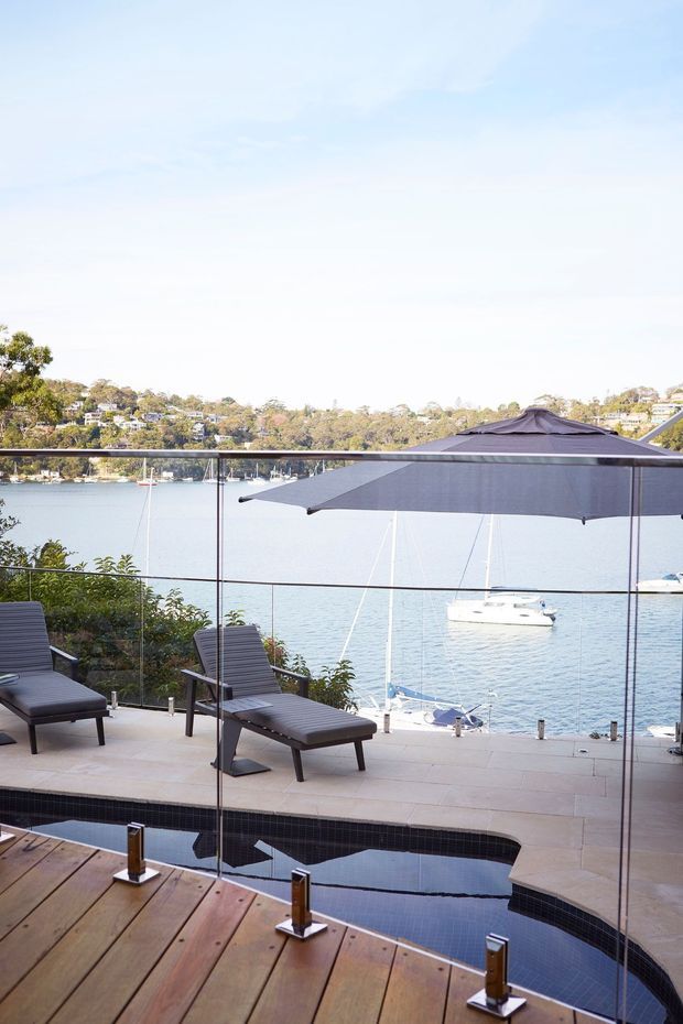 A bright waterfront terrace overlooking a calm bay, with a narrow swimming pool in the foreground, timber decking, pale stone paving, frameless glass balustrades, dark reclining loungers, and a large dark outdoor umbrella. Boats and hillside homes are visible across the water.
