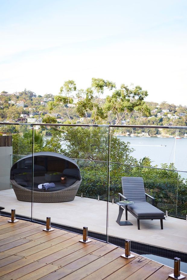 A sunlit waterfront terrace overlooking a calm inlet, with a glass balustrade, timber decking, pale tiled paving, a rounded woven outdoor daybed, and a reclining lounge chair. Established vegetation and hillside residences are visible across the water.