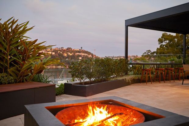 A contemporary outdoor terrace overlooking a waterfront, with a built-in rectangular fire pit in the foreground, large dark raised planters, lush screening vegetation, and a covered outdoor seating area with a dining table and stools. The setting appears elevated above a body of water with a suburban hillside beyond.