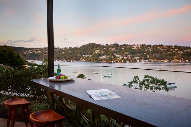 A waterfront terrace or balcony with a long stone-clad outdoor counter overlooking a calm harbour. Two timber stools, potted greenery, a fruit platter, and design magazines create a relaxed entertaining and work setting, with boats and hillside homes visible across the water.