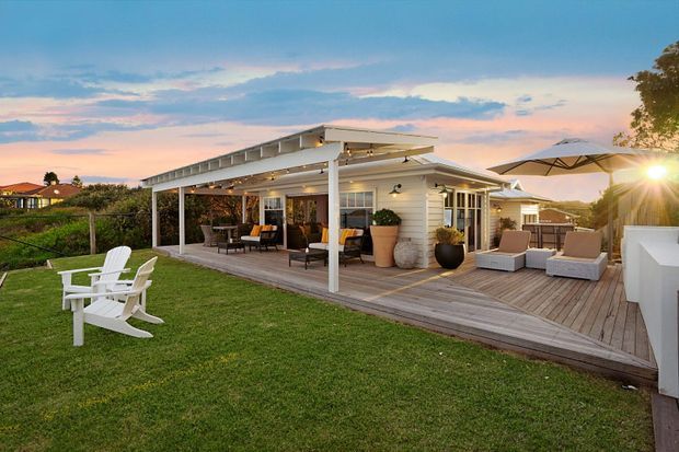 A single-storey coastal-style house with a broad covered outdoor entertaining area and timber deck overlooking a manicured lawn. The deck includes lounge seating, dining furniture, potted plants, umbrellas and built-in outdoor cabinetry, with warm evening lighting and a suburban coastal setting.