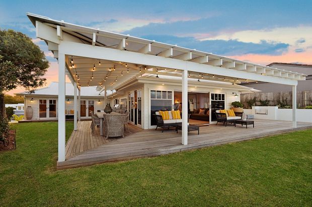 A contemporary single-storey home with an expansive covered outdoor living area and timber decking. White posts and exposed roof framing create a generous pergola, with string lights, an outdoor dining setting, lounge seating, and glazed doors connecting to the interior. The home sits beside a broad lawn in a suburban or landscaped setting, photographed at dusk.