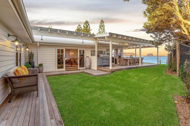A single-storey coastal home arranged around a manicured lawn and covered outdoor entertaining area. The exterior features light horizontal weatherboard cladding, timber decking, a broad pergola with exposed beams, outdoor dining and lounge furniture, and warm festoon-style lighting. Glimpses of blue water and a distant headland are visible beyond the patio.