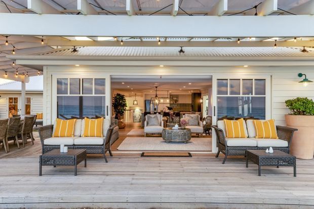 A bright coastal-style covered outdoor entertaining area with a wide timber deck, two lounge seating arrangements, striped yellow-and-white cushions, ceiling fans, festoon lighting, and a large opening into an adjoining living room. Ocean views are visible through the side windows, with white horizontal cladding and exposed roof structure.