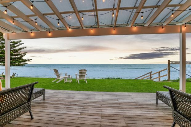 A covered outdoor entertaining area with a timber deck, exposed timber pergola framing, warm festoon lighting, and wicker seating overlooking a manicured lawn and the ocean. The open coastal outlook and relaxed outdoor furniture create a serene seaside setting.