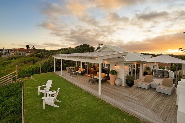 A single-storey coastal-style home with a broad covered verandah and expansive timber decking overlooking a landscaped lawn and garden. The outdoor entertaining area includes lounge seating, an outdoor dining setting, umbrellas, potted plants and white Adirondack chairs. The house has light weatherboard cladding, white trims, pitched gabled roof sections and large glazed doors, photographed at sunset.