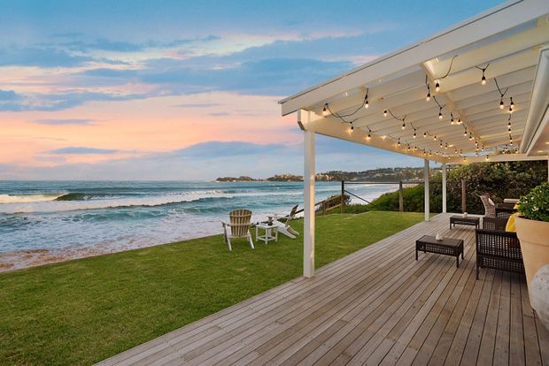 A seaside outdoor entertaining area with a broad timber deck, covered pergola, and overhead festoon lighting. Outdoor lounge furniture and potted greenery sit beside a lawn overlooking waves, sandy shoreline, and a rocky coastal headland at sunset.