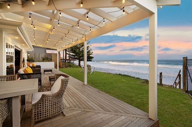 A spacious covered beachfront deck overlooking the ocean, furnished with a dining table, woven outdoor chairs and a lounge area. White-painted posts and beams support a pergola-style roof strung with festoon lights, while timber decking extends toward a grassed shoreline and stair access to the beach. The scene is warmly lit at sunset with a relaxed coastal atmosphere.
