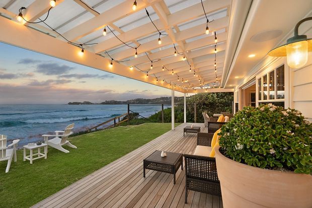 A coastal outdoor entertaining area overlooking the ocean, featuring a covered timber deck with a white exposed-beam ceiling, hanging string lights, wicker lounge furniture, built-in glazing and planters. A manicured lawn with Adirondack-style chairs extends toward the rocky shoreline.
