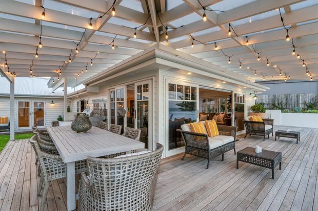 A spacious covered outdoor entertaining area with a large timber deck, dining table and woven chairs in the foreground, plus a lounge setting beside a glazed white-clad pavilion. The space features extensive glass doors and windows, exposed timber framing, a translucent roof, and warm string lighting. It opens onto a smaller courtyard with additional seating and landscaping, creating a bright, inviting indoor-outdoor living environment.