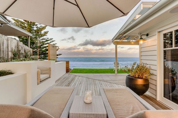 A coastal outdoor entertaining area overlooking the ocean, with a timber deck, outdoor lounge furniture, large umbrella, built-in rendered seating, and a light weatherboard-clad building with covered patio lighting. The setting has a relaxed beachfront character and open sea views.