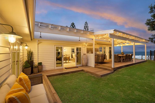 Twilight exterior view of a coastal-style home with white weatherboard cladding, a covered timber deck and pergola, outdoor dining area, built-in outdoor kitchen or bar, and a lounge seating area overlooking the ocean. Warm integrated lighting creates an inviting outdoor entertaining setting.