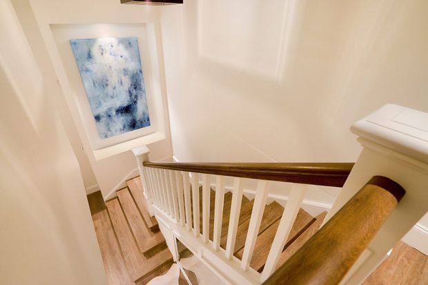 A bright residential staircase viewed from the upper landing, with white painted balustrades and newel posts, a polished dark timber handrail, and warm timber stair treads. White paneled walls and an abstract blue artwork add subtle detail.