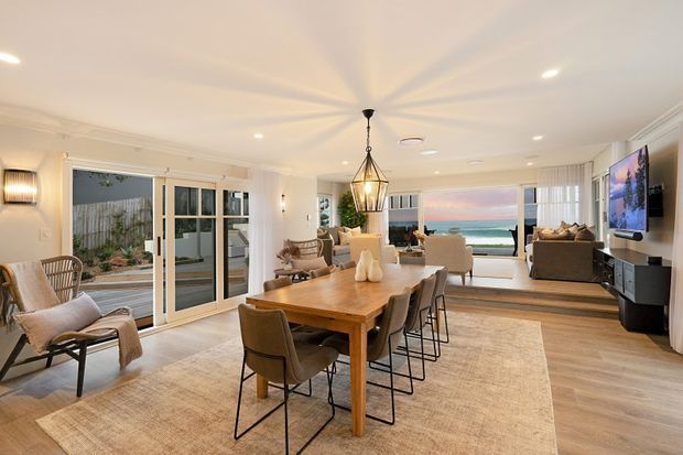 A bright, open-plan coastal living and dining space with a large timber dining table, upholstered dining chairs, lounge seating, woven armchairs, and a large neutral rug. White walls and ceiling, pale timber flooring, extensive glazing, and sliding doors create an airy connection to the outdoor courtyard and ocean view. A central pendant light and recessed ceiling lights provide illumination, while a wall-mounted television is visible in the living area.