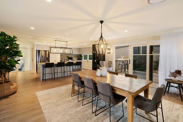 Bright, open-plan contemporary dining and kitchen interior with a large timber dining table, upholstered dining chairs, pendant lighting, light timber flooring, and a generous kitchen island with bar seating. Full-height glazed doors and sheer curtains open toward an outdoor area, while warm neutral finishes and indoor planting create an inviting atmosphere.